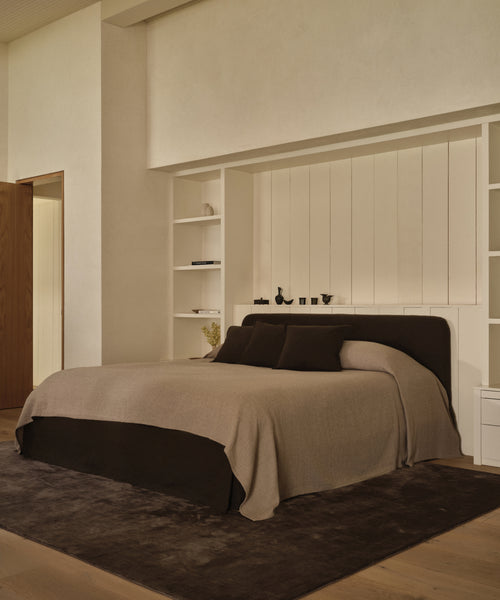 A modern, minimalist bedroom features the Cove Bed with an upholstered headboard, beige and brown Libeco linen bedding, matching pillows, built-in white shelves, light walls, wood flooring, a dark rug, and subtle decor accents.