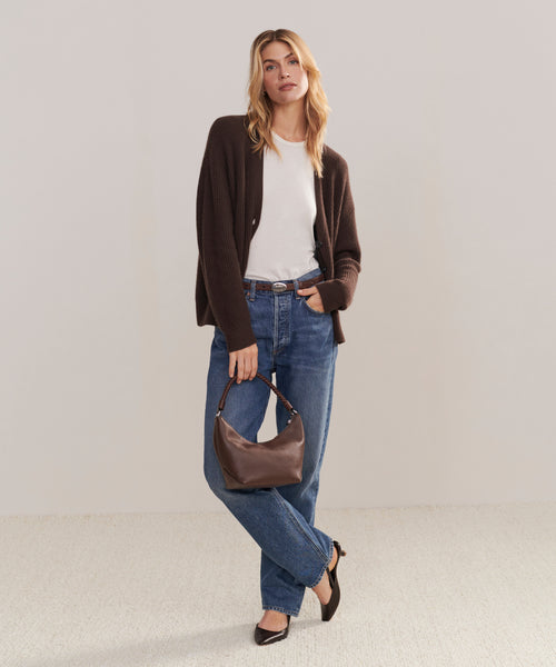 A woman stands against a plain background wearing a dark brown cardigan, white top, blue jeans, and black flats. She holds the Crescent Shoulder Bag and poses casually with one leg crossed in front of the other.
