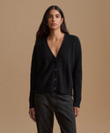 A woman modeling a black ribbed knit cardigan with a V-neck and front buttons, paired with dark gray jeans, against a solid tan studio background.