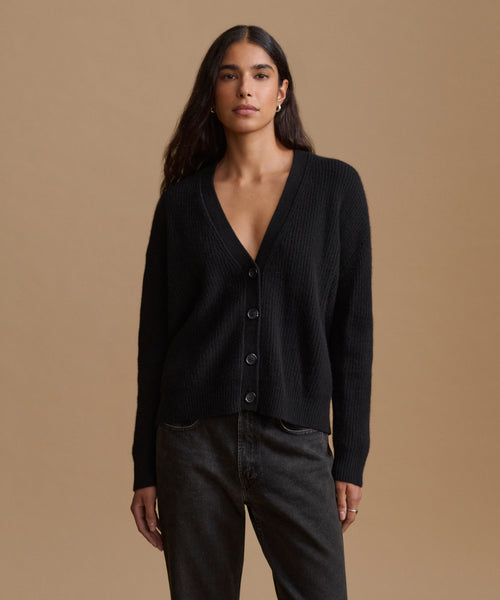 A woman modeling a black ribbed knit cardigan with a V-neck and front buttons, paired with dark gray jeans, against a solid tan studio background.