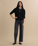 A woman with long, wavy hair stands against a beige background wearing the Cropped Cashmere Cocoon Cardigan in black, dark wide-leg jeans, black flats, and layered necklaces. She rests one hand on her hip.