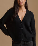 A woman wearing a black ribbed knit cardigan sweater with a V-neck and dark buttons, paired with dark jeans, against a solid warm beige background.