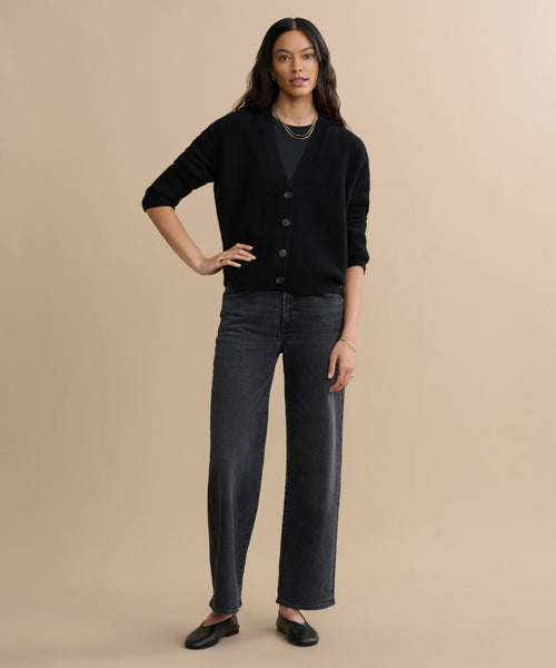 A woman with long, wavy hair stands against a beige background wearing the Cropped Cashmere Cocoon Cardigan in black, dark wide-leg jeans, black flats, and layered necklaces. She rests one hand on her hip.