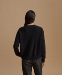 A model viewed from behind wearing a black ribbed knit sweater and dark gray jeans, set against a solid tan studio background.