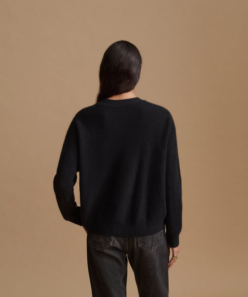 A model viewed from behind wearing a black ribbed knit sweater and dark gray jeans, set against a solid tan studio background.