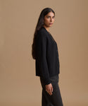 A woman with long dark hair is shown in profile wearing a black ribbed knit cardigan and dark trousers, standing against a solid warm beige studio background.