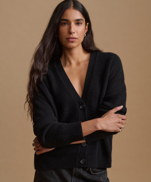 A woman wearing a black ribbed knit cardigan with a V-neck and dark buttons, posed against a solid warm beige studio background.