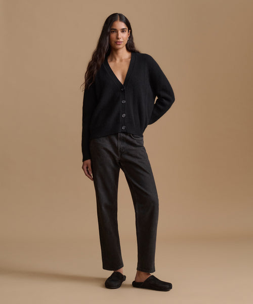 A woman modeling a black button-front cardigan, dark charcoal gray straight-leg jeans, and black slip-on shoes against a solid beige studio background.