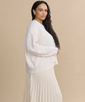 A woman with long, wavy dark hair stands in profile against a beige background, wearing an ivory Cropped Cashmere Cocoon Cardigan and a matching pleated skirt.