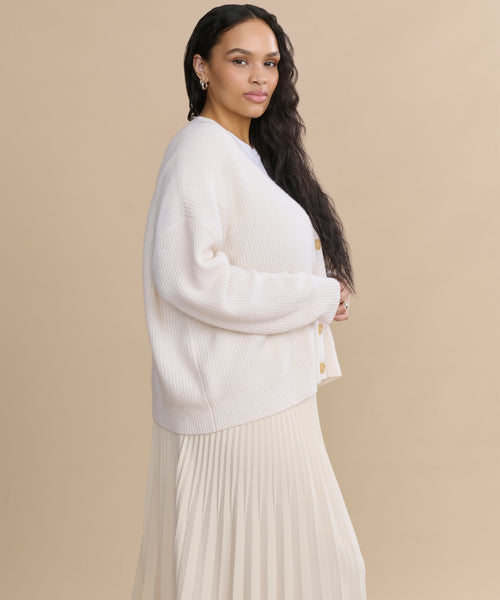 A woman with long, wavy dark hair stands in profile against a beige background, wearing an ivory Cropped Cashmere Cocoon Cardigan and a matching pleated skirt.