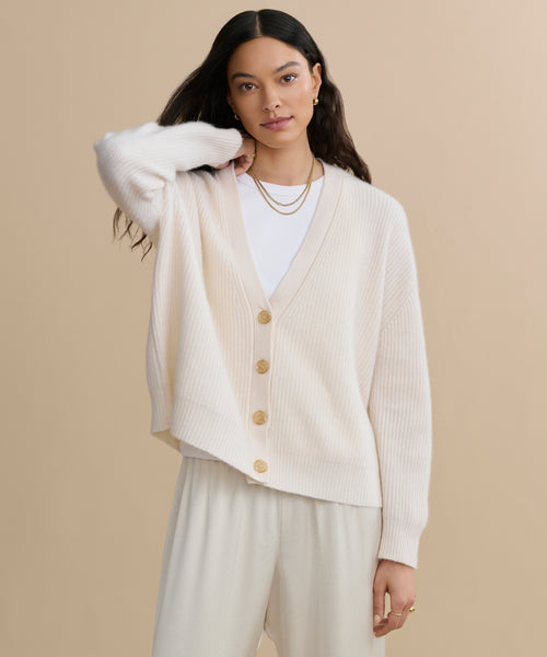 A woman with long dark hair wears the Cropped Cashmere Cocoon Cardigan in ivory over a white shirt and cream pants, accessorized with gold hoop earrings and layered necklaces, standing against a beige background.