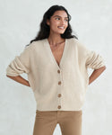 Woman with long dark hair wears the Cropped Cashmere Cocoon Cardigan with brown buttons and tan pants, standing hands on hips and smiling against a light background.