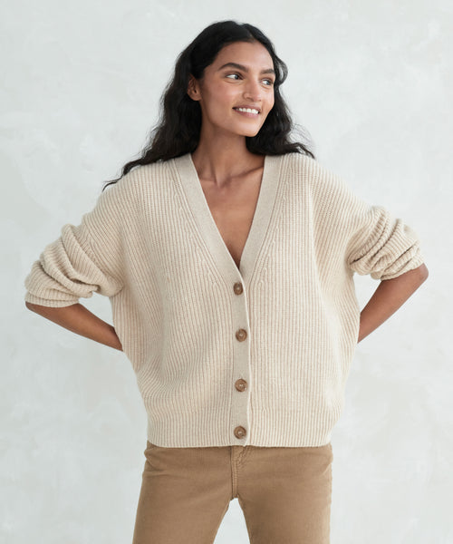 Woman with long dark hair wears the Cropped Cashmere Cocoon Cardigan with brown buttons and tan pants, standing hands on hips and smiling against a light background.