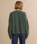 A person with short blonde hair, wearing the Cropped Cashmere Cocoon Cardigan and blue jeans, stands facing away from the camera against a plain beige background.