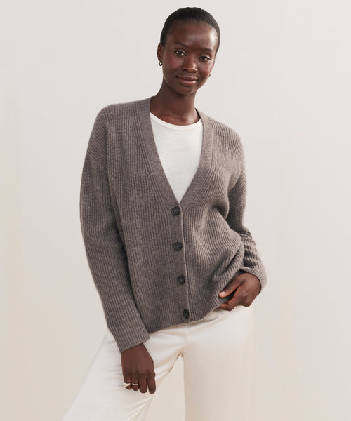 Wearing the Cropped Cashmere Cocoon Cardigan over a white top and pants, a person stands against a plain light background, smiling slightly with one hand in their pocket.