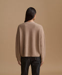 A model viewed from the back wearing a beige ribbed knit sweater and dark jeans, set against a solid warm beige studio background.