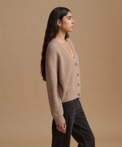 A woman in a side profile wearing a beige ribbed knit cardigan with dark jeans, set against a solid warm beige studio background.