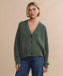 A person wears the Cropped Cashmere Cocoon Cardigan in olive green with a V-neck and blue jeans, standing against a plain beige background.