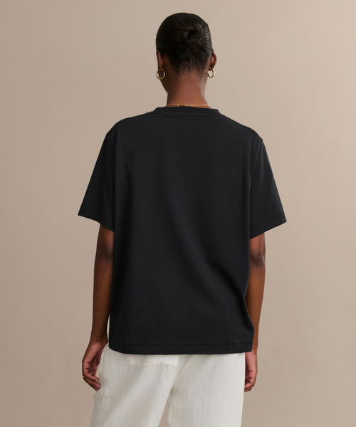 A person with short hair in a bun faces away from the camera, wearing a relaxed fit Crosby Tee and white pants, both made from premium jersey cotton, set against a neutral beige background.