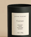 The Currant Glass Candle is a hand-poured 10 oz glass candle with a white label featuring notes of Bergamot, Cassis, White Rose, and Oakmoss. Made from natural coconut soy wax, it sits elegantly against a beige backdrop.