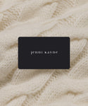 A black Jenni Kayne eGift card with white lowercase lettering rests on a soft, cream-colored knitted fabric background, making it the perfect effortless gift.