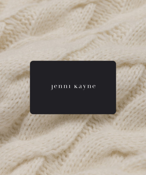 A black Jenni Kayne eGift card with white lowercase lettering rests on a soft, cream-colored knitted fabric background, making it the perfect effortless gift.