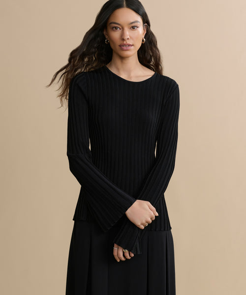 Against a beige background, a woman with long, wavy dark hair wears the Dara Top—a black ribbed piece with subtle bell sleeves—paired with a black skirt. Her arms are crossed loosely in front of her.