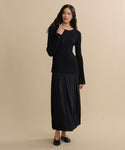 A woman wearing the Dara Top and a long black skirt stands against a beige background. She has long wavy hair, is wearing black flats, and has one hand touching her neck.
