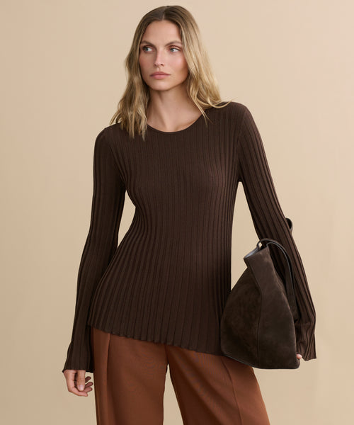 A woman with wavy blonde hair wears the fitted, long-sleeve brown Dara Top and loose brown pants, holding a dark brown handbag against a beige background.