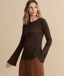 A woman with wavy, shoulder-length blonde hair wears the Dara Top and brown pants, standing against a plain beige background with one hand in her pocket, looking to the side.