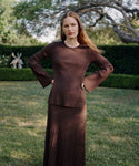 A woman with long hair stands on grass wearing the Dara Top and a matching A-line skirt, looking at the camera with a neutral expression, trees and hedges in the background.