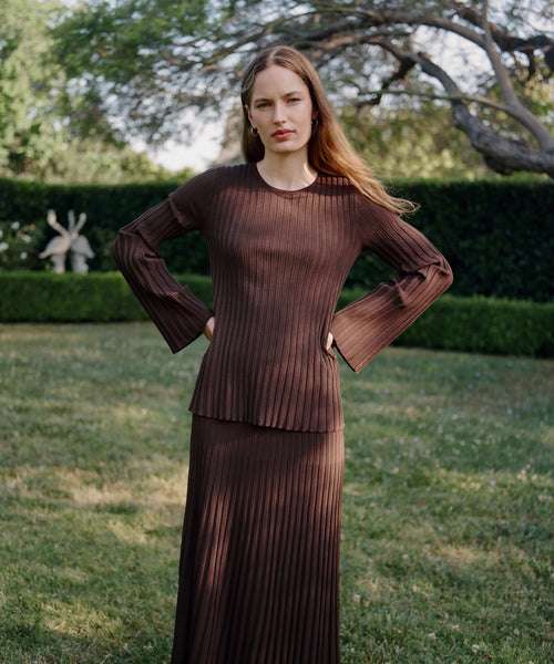 A woman with long hair stands on grass wearing the Dara Top and a matching A-line skirt, looking at the camera with a neutral expression, trees and hedges in the background.