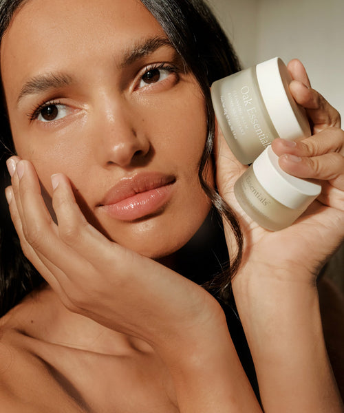 A woman with glowing skin gently touches her face while holding two jars of Oak Essentials, including their Cleansing Balm, as she poses in natural light.
