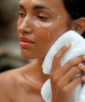 A woman with dewy skin gently presses a white towel to her cheek, looking relaxed and refreshed—likely after using Cleansing Balm in her skincare routine.