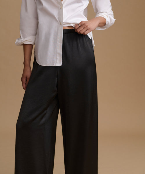 A model wearing black wide-leg trousers and a white button-down shirt, shown from the chest down against a solid beige background.
