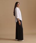 A woman stands in profile wearing a loose white long-sleeve blouse with rolled cuffs and black wide-leg trousers, photographed against a plain warm beige studio background.