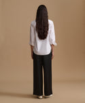 A woman viewed from behind wearing a white long-sleeve blouse with ruffled cuffs and black wide-leg trousers, standing against a plain beige studio background.