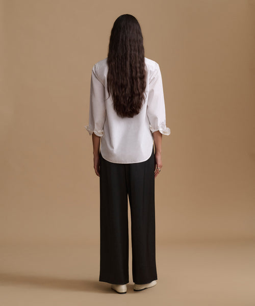 A woman viewed from behind wearing a white long-sleeve blouse with ruffled cuffs and black wide-leg trousers, standing against a plain beige studio background.
