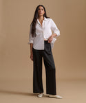 A woman modeling a white long-sleeve button-up shirt and black wide-leg trousers, paired with white pointed flats, against a solid beige studio backdrop.