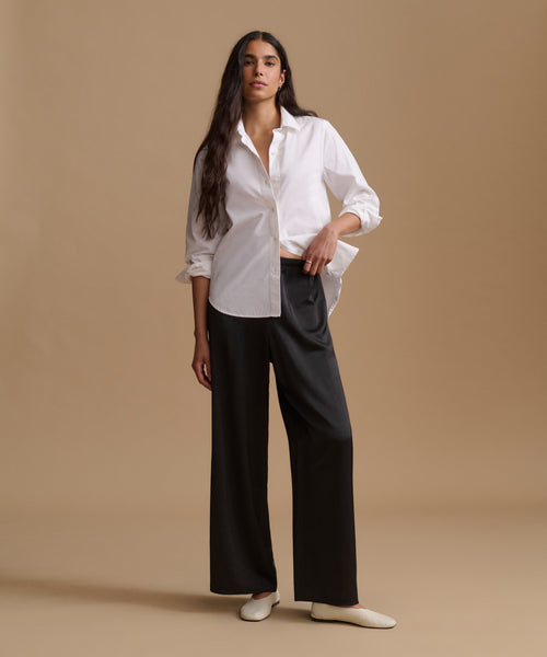 A woman modeling a white long-sleeve button-up shirt and black wide-leg trousers, paired with white pointed flats, against a solid beige studio backdrop.