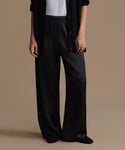 A model wearing black wide-leg satin trousers, styled with a white top, a black button-up cardigan, and black pointed-toe flats, set against a solid beige studio background.