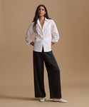 A woman stands against a warm beige studio background wearing a white long-sleeve button-up shirt, black wide-leg pants, and white flats.