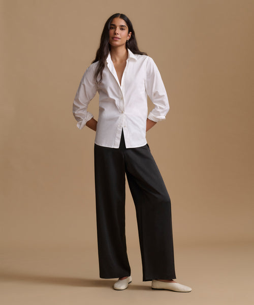A woman stands against a warm beige studio background wearing a white long-sleeve button-up shirt, black wide-leg pants, and white flats.