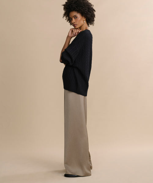 A woman stands in profile against a beige background, wearing the Demi Pant and a loose black sweater. She looks thoughtfully at the camera with one hand touching her chin, her curly hair styled naturally for a sophisticated, flowy look.