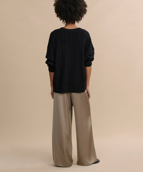 A person with short curly hair stands relaxed, facing away, wearing luxe pull-on Demi Pant and a loose black sweater against a plain beige background.