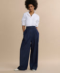 A person with curly hair wears a white button-up shirt, Denim Newport Trouser in dark blue, and white pointed shoes, standing against a plain beige background.