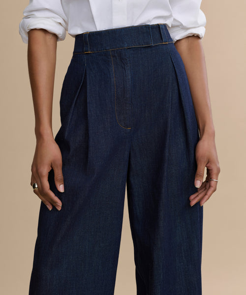 Wearing the Denim Newport Trouser—high-waisted, straight leg dark denim—paired with a white long-sleeve shirt and rolled sleeves, standing before a beige background, this look is an effortless tailored essential.