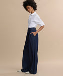 A person stands sideways against a beige background, wearing a white collared shirt with rolled-up sleeves, Denim Newport Trouser with a straight leg, and white shoes. Their hands are in their pockets and they have short curly hair.
