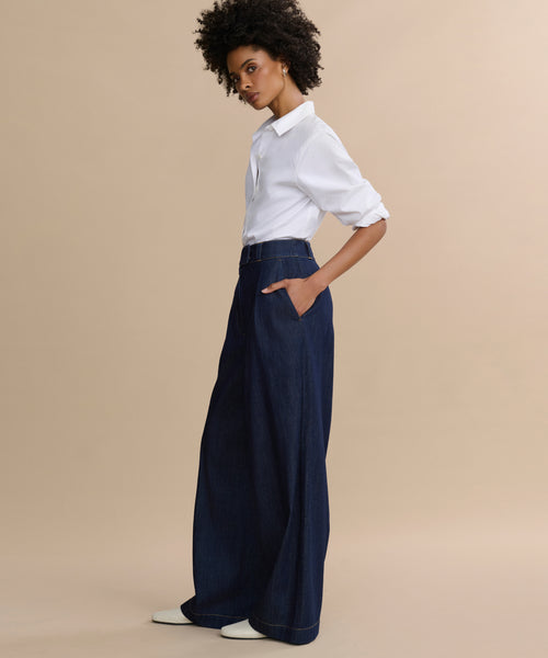 A person stands sideways against a beige background, wearing a white collared shirt with rolled-up sleeves, Denim Newport Trouser with a straight leg, and white shoes. Their hands are in their pockets and they have short curly hair.
