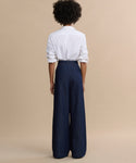 A person stands facing away, wearing a white long-sleeve shirt with rolled-up sleeves and Denim Newport Trousers featuring a high-waisted, straight-leg fit, set against a plain beige background.